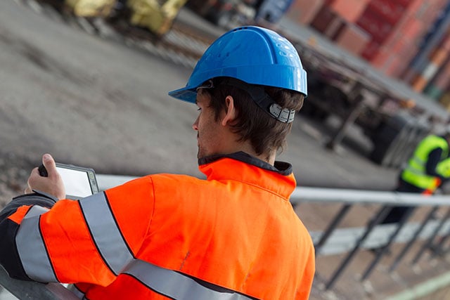 Professional in a hard hat uses a tablet at a harbour area.