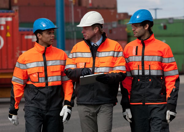 Three people in hard hats, walking and discussing. Containers on the background.